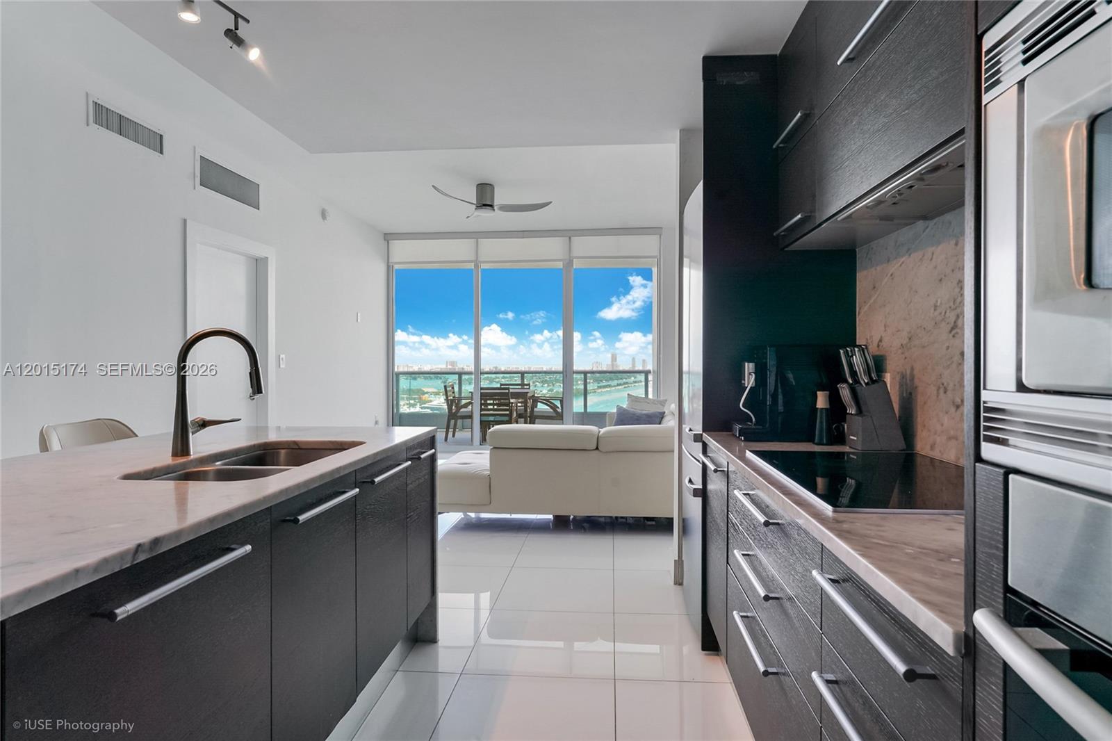 A modern kitchen with dark cabinetry and stone countertops overlooks a living area and balcony — 900 Biscayne Blvd # 3407, Miami FL 33132, MIAMI, FL