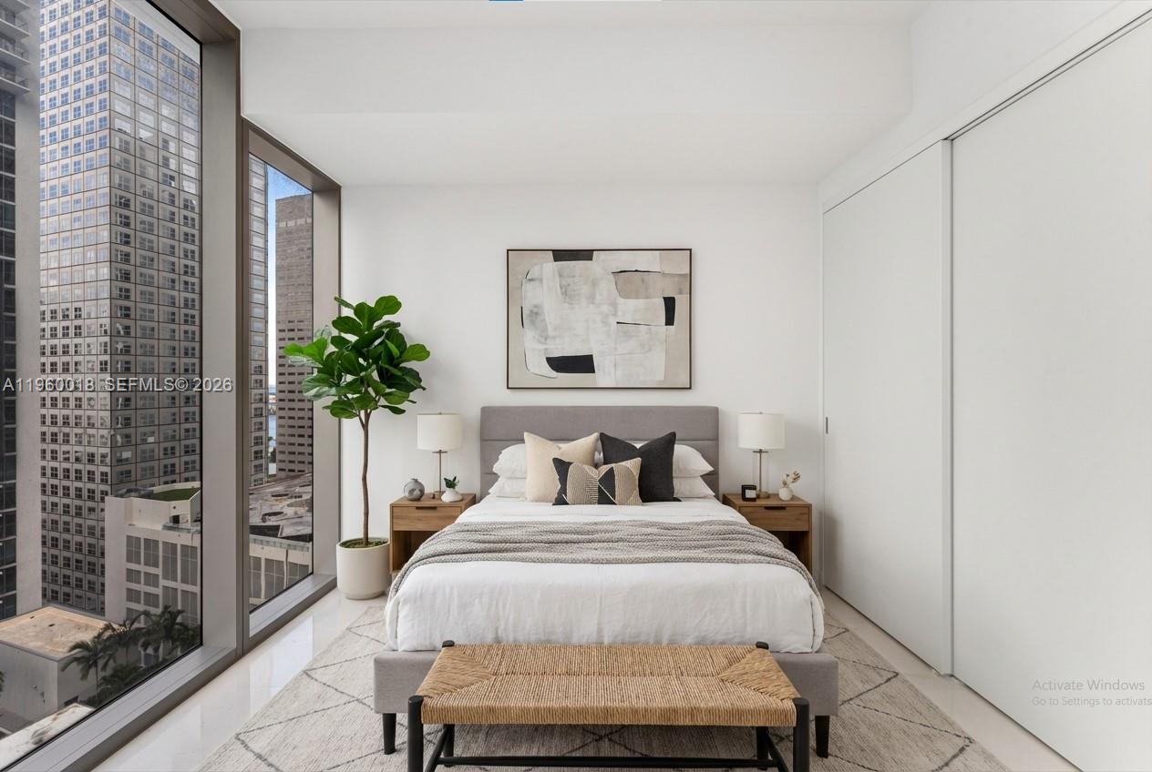 A modern bedroom features a bed, nightstands, a plant, and floor-to-ceiling windows overlooking city buildings — 300 BISCAYNE BLVD WAY # 2009, Miami FL 33131, MIAMI, FL