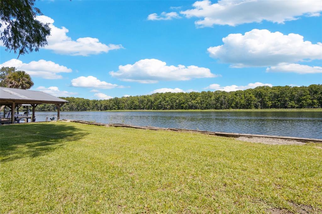 A grassy backyard overlooks a lake with a wooden boat dock under a metal roof — 15471 NW 46TH LANE, CHIEFLAND, FL