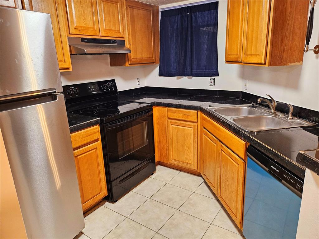 A kitchen features oak cabinets, black countertops, a stainless steel refrigerator, and tile flooring — 1916 LAKE ATRIUMS CIRCLE UNIT 25, ORLANDO, FL