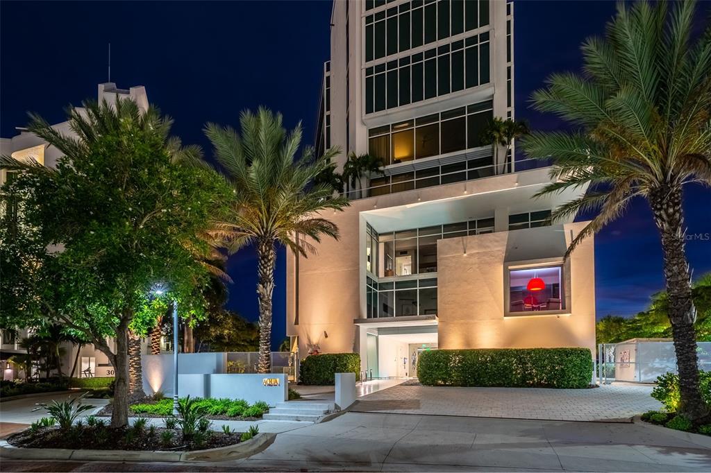 The exterior of a modern high-rise building at night features palm trees and landscaping — 280 GOLDEN GATE POINT UNIT 300, SARASOTA, FL