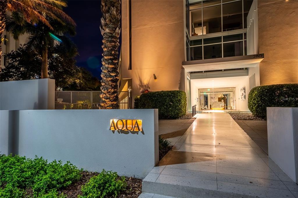 The exterior entrance of a modern building features a lit sign reading Aqua at night — 280 GOLDEN GATE POINT UNIT 300, SARASOTA, FL