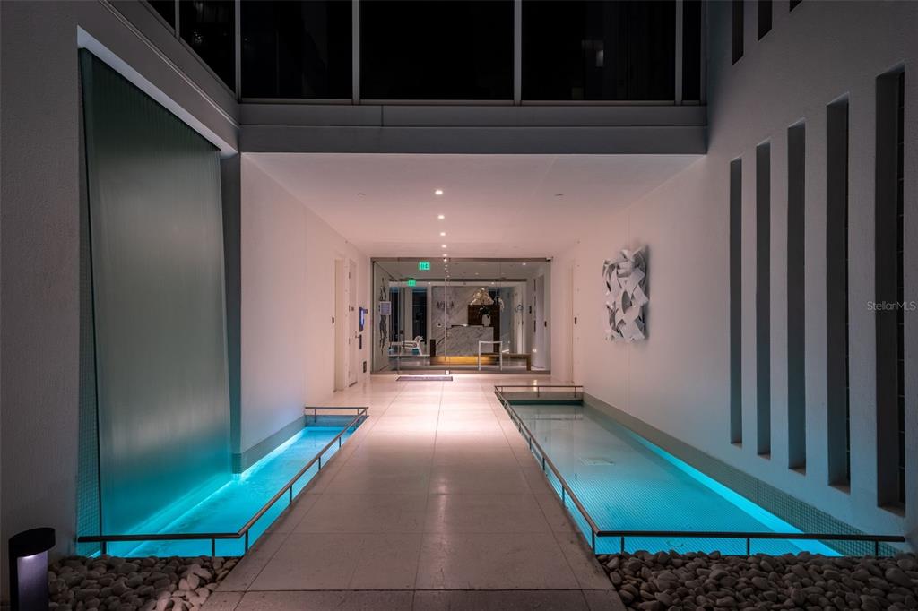 A modern building entrance features a central walkway flanked by illuminated indoor water features — 280 GOLDEN GATE POINT UNIT 300, SARASOTA, FL