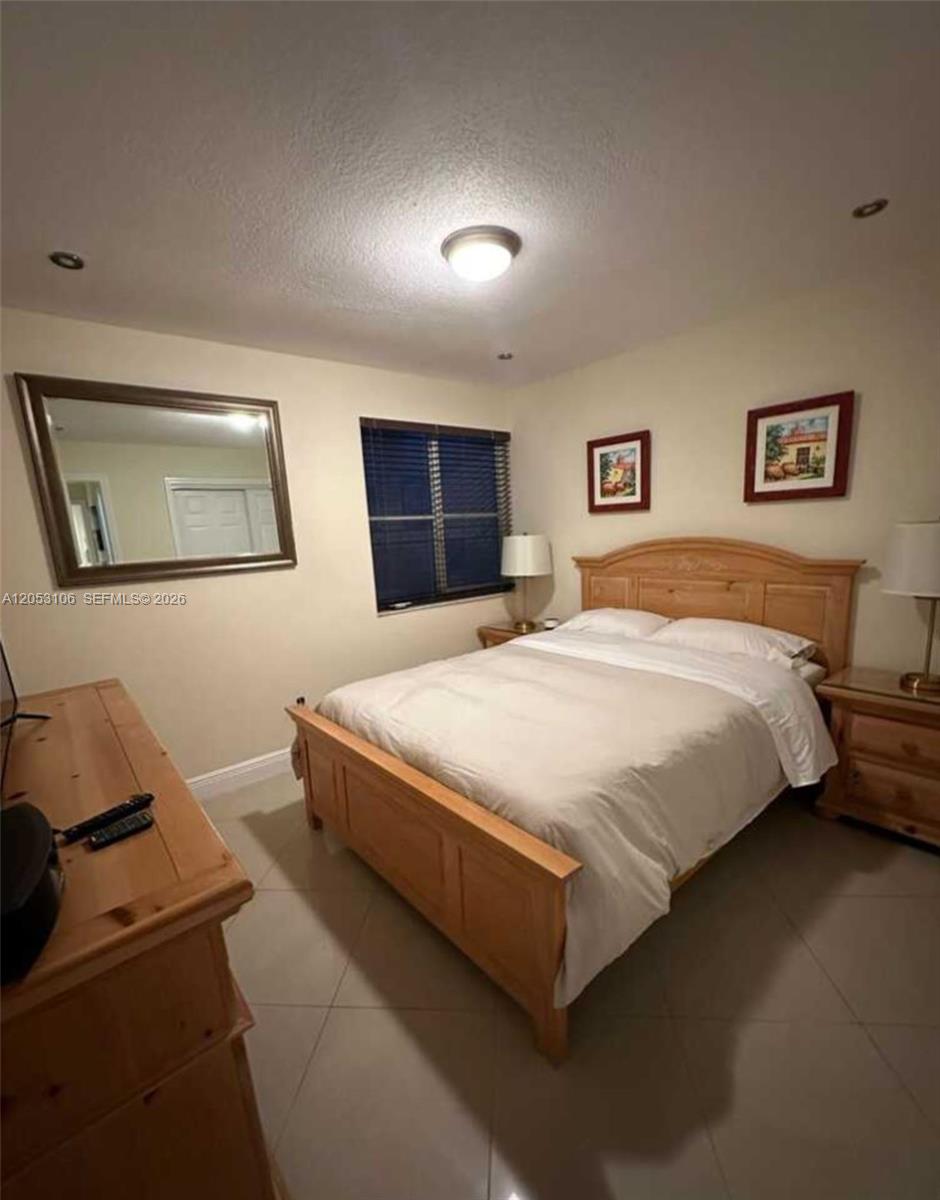 A bedroom features a wooden bed frame, a dresser, a mirror, and two framed pictures — MIAMI, FL