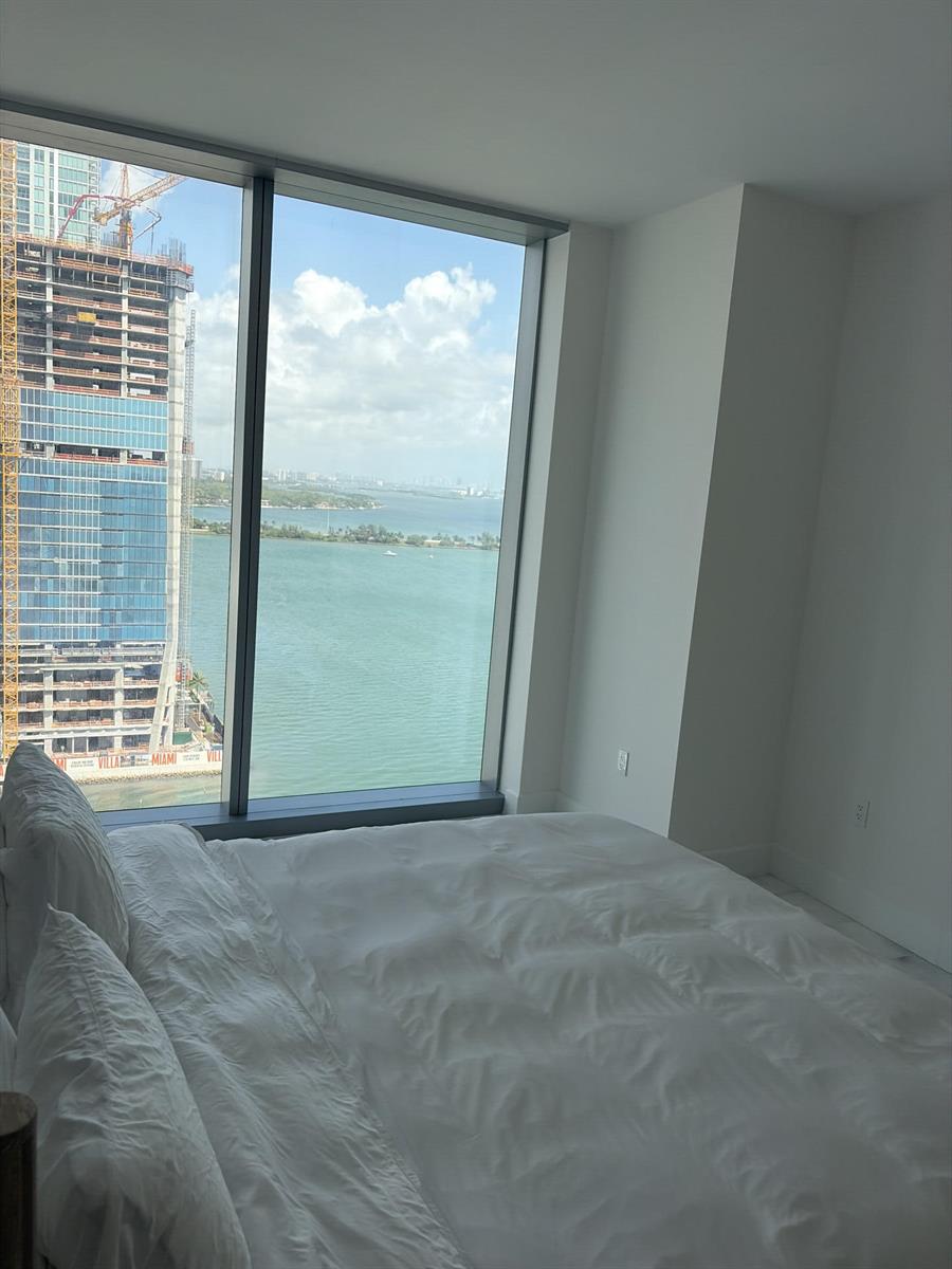 A bedroom with a white bed faces floor-to-ceiling windows overlooking a bay and construction site — 700 NE 26th Terrace # 2006, Miami FL 33137, MIAMI, FL