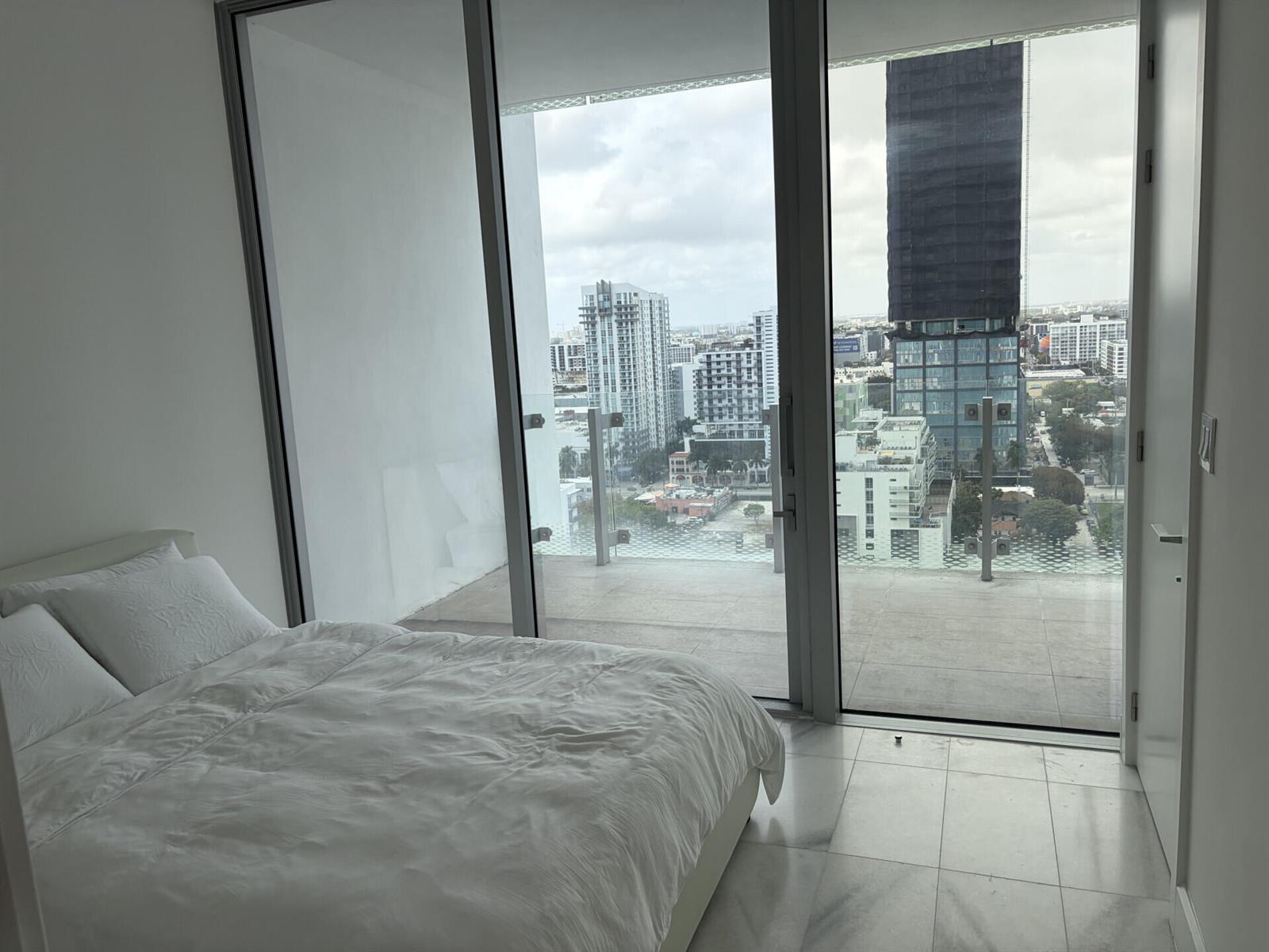 A bedroom with white bedding and marble floors features floor-to-ceiling windows overlooking a city skyline — 700 NE 26th Terrace # 2006, Miami FL 33137, MIAMI, FL