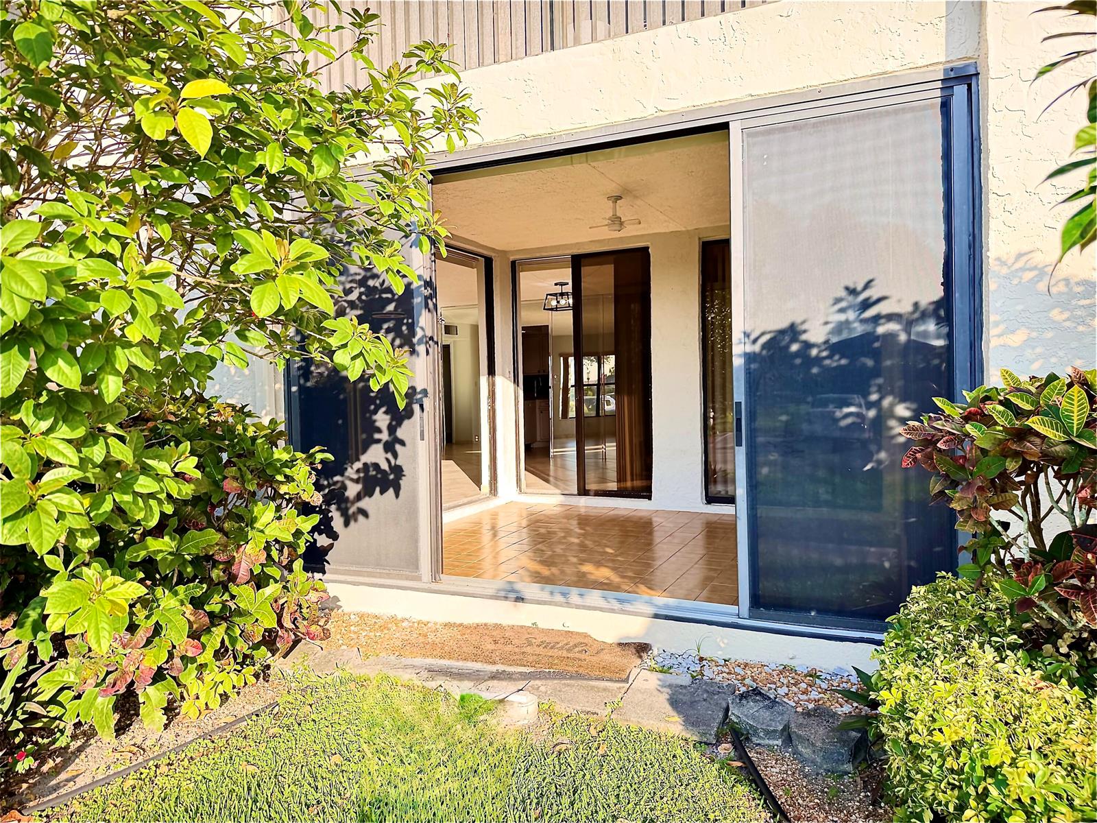 A tiled patio with sliding glass doors is framed by lush green bushes and landscaping — 8657 Boca Glades Boulevard W # C, Boca Raton FL 33434, BOCA RATON, FL