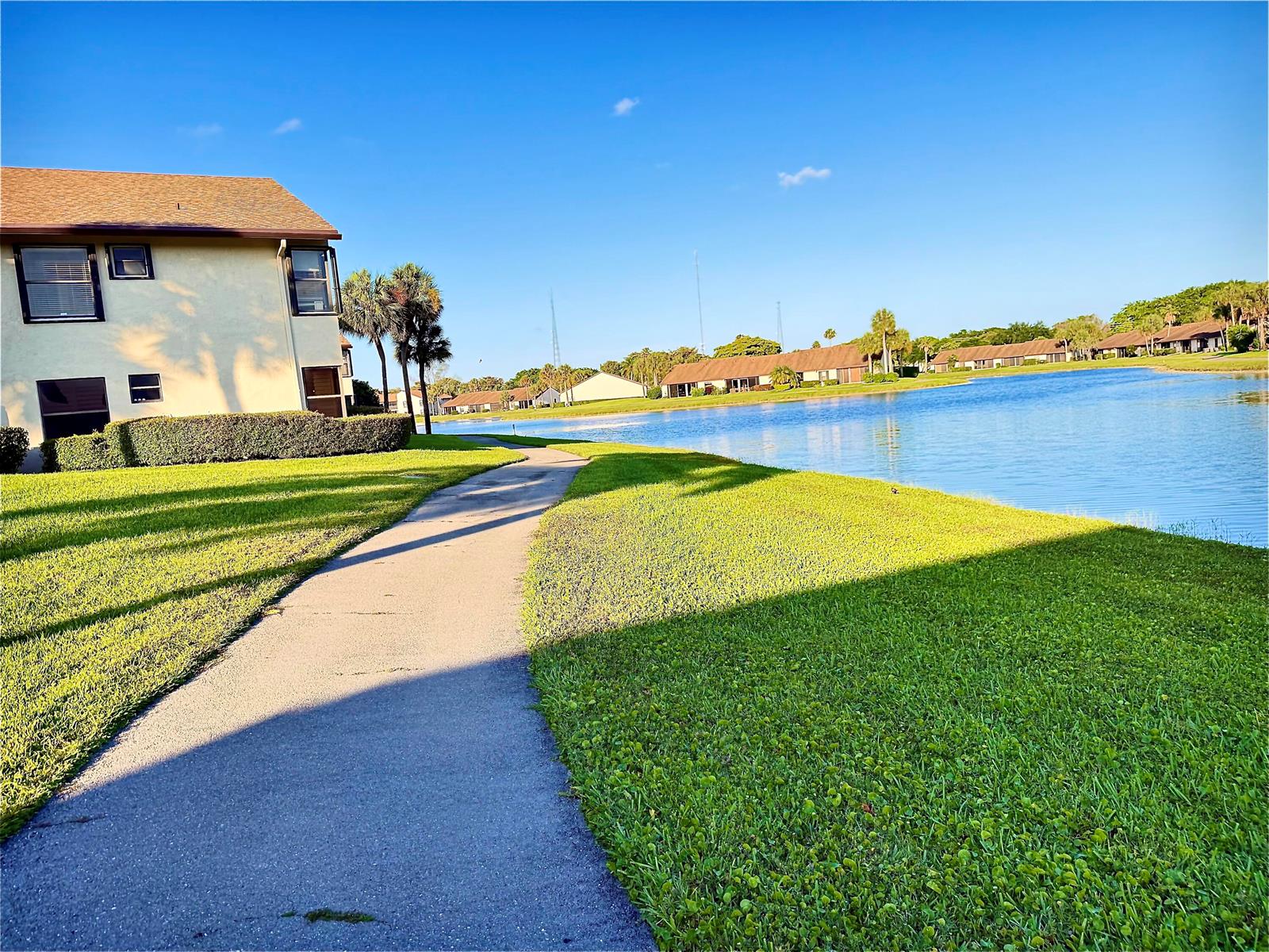 A paved walkway winds past a multi-story building and a large pond under a clear sky — 8657 Boca Glades Boulevard W # C, Boca Raton FL 33434, BOCA RATON, FL