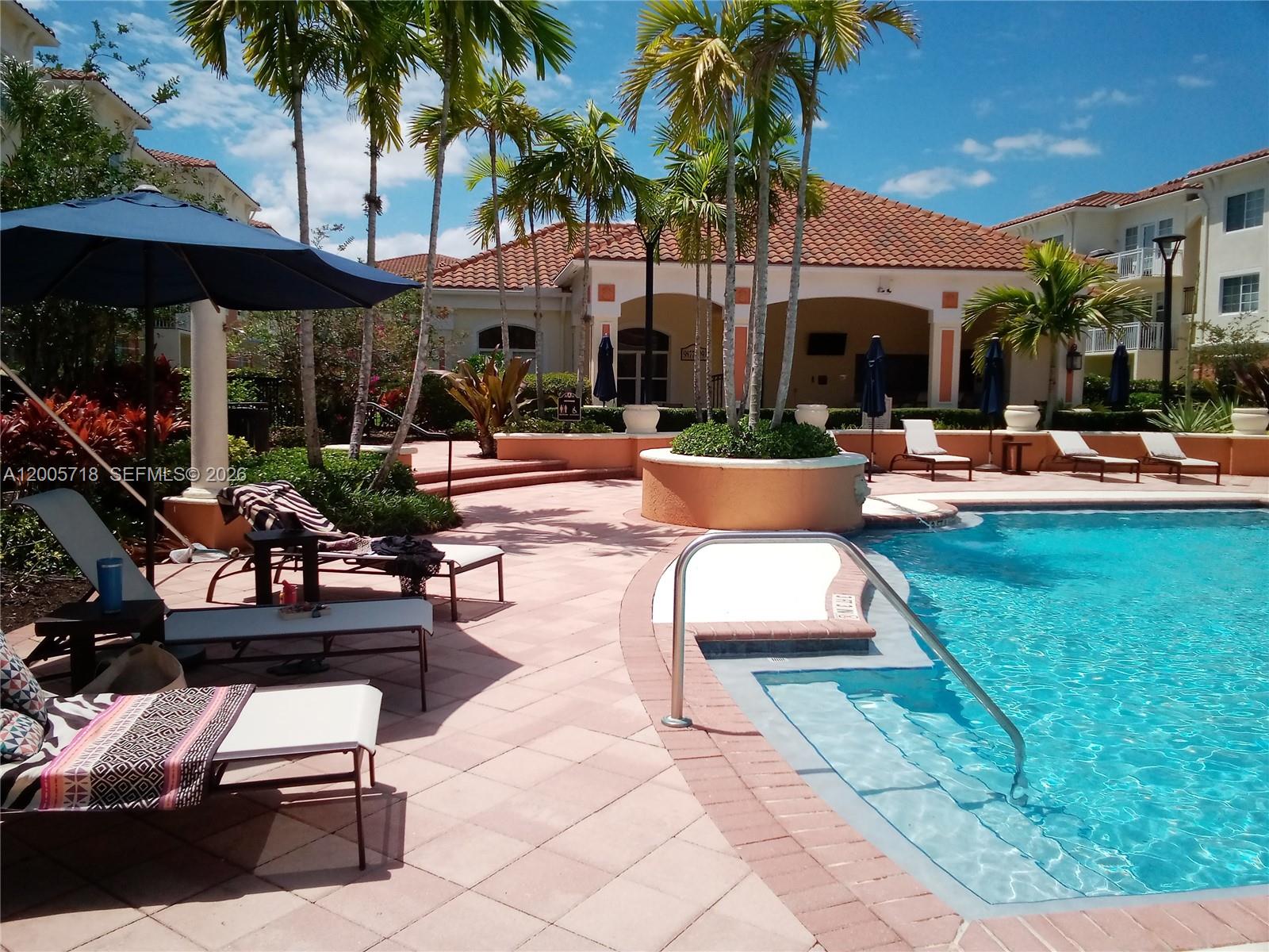 A swimming pool with lounge chairs and palm trees sits in front of a clubhouse — 9845 Baywinds Dr # 6308, West Palm Beach FL 33411, WEST PALM BEACH, FL