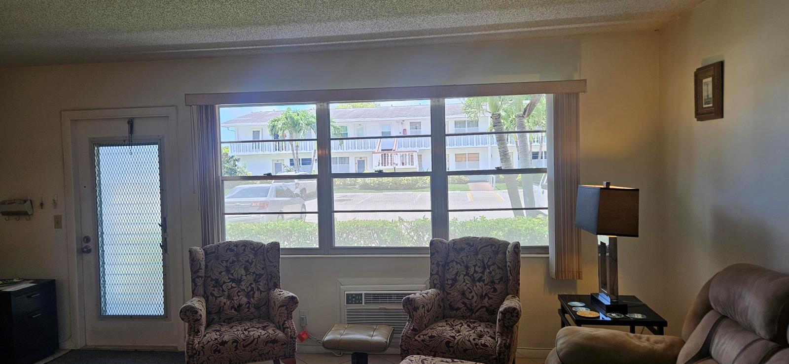 A living room features two patterned armchairs, a window view of an apartment building, and furniture — 27 Newport B, Deerfield Beach FL 33442, DEERFIELD BEACH, FL