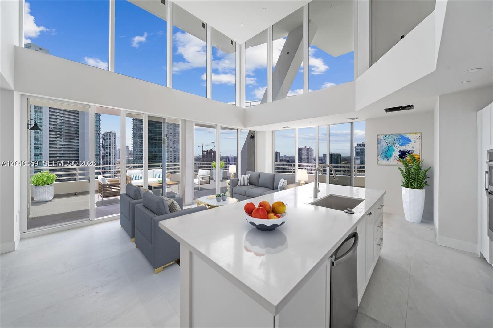A modern kitchen island with a fruit bowl overlooks a living area with floor-to-ceiling windows — 555 NE 34th St, Miami FL 33137, MIAMI, FL