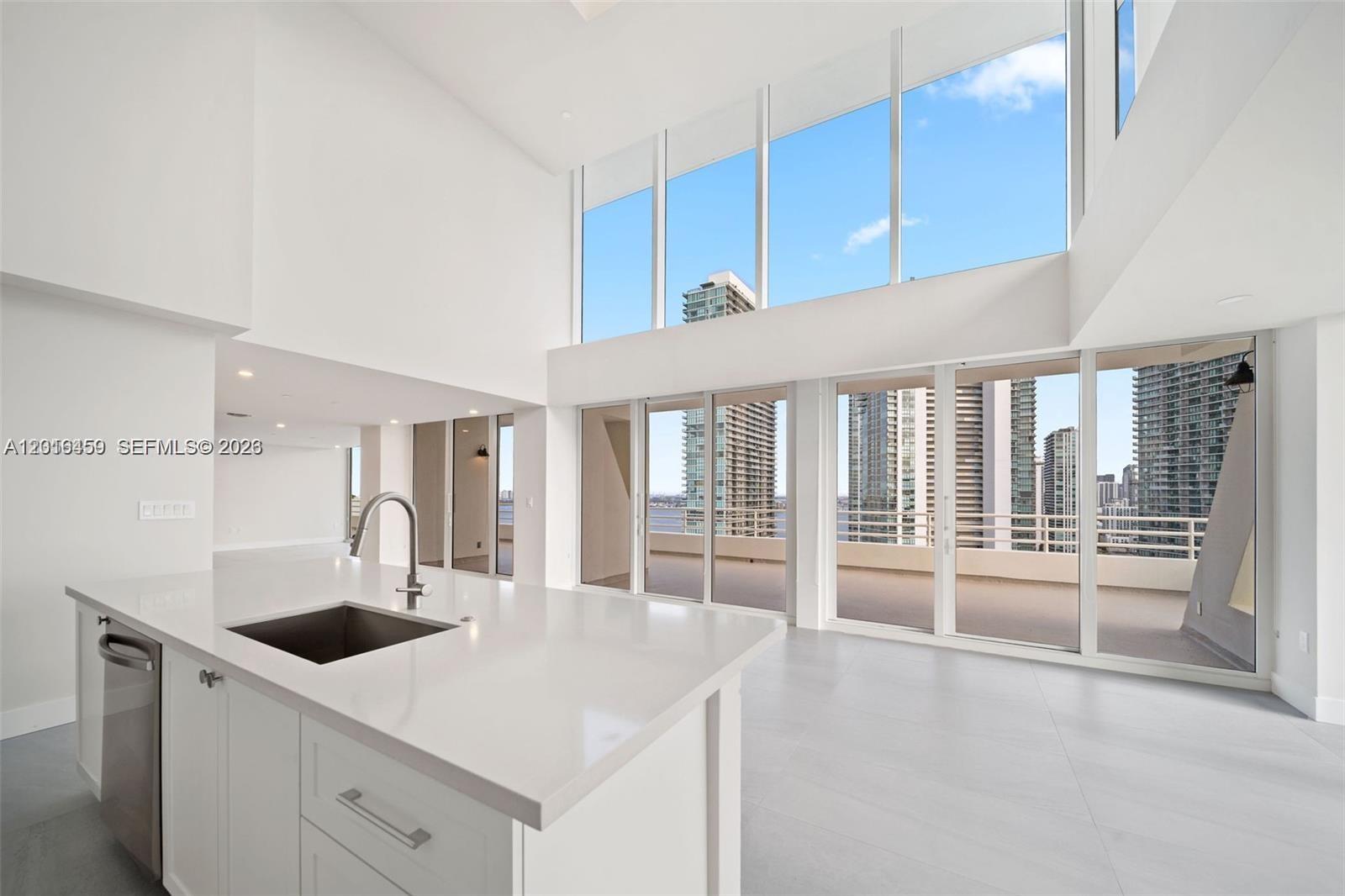 A modern kitchen with a white island overlooks a double-height living area with floor-to-ceiling windows — 555 NE 34th St, Miami FL 33137, MIAMI, FL
