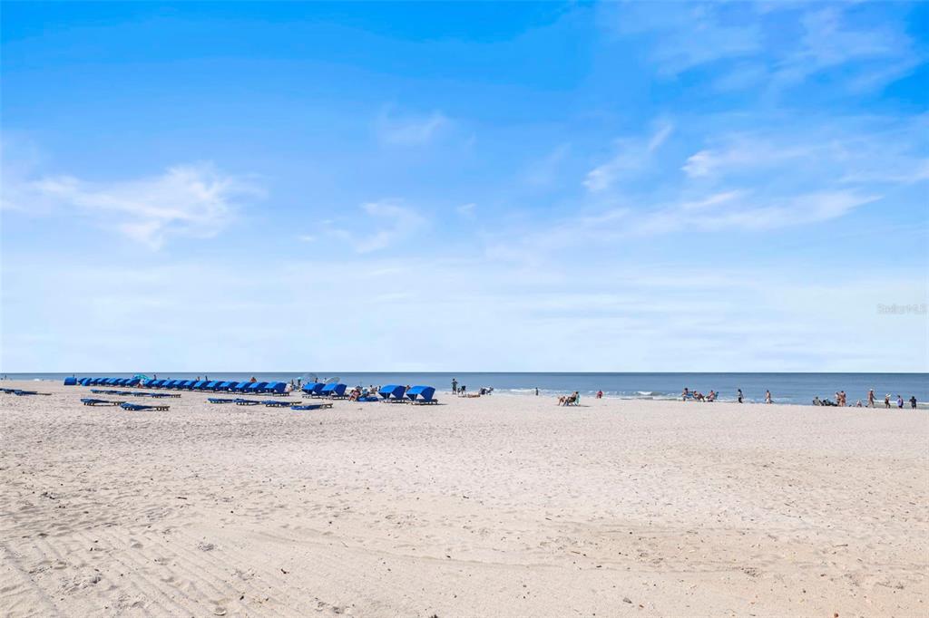 A wide, sandy beach features rows of blue umbrellas and lounge chairs under a blue sky — 20040 GULF BOULEVARD UNIT 302, INDIAN SHORES, FL