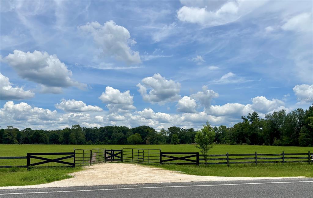 A gravel driveway leads to a metal gate opening into a large, fenced grassy pasture — NW 202ND STREET, HIGH SPRINGS, FL