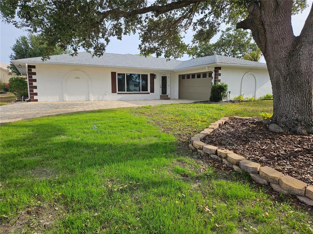 A single-story white house with a paved driveway, a two-car garage, and a large tree — 1508 FLAMINGO LANE, SUN CITY CENTER, FL