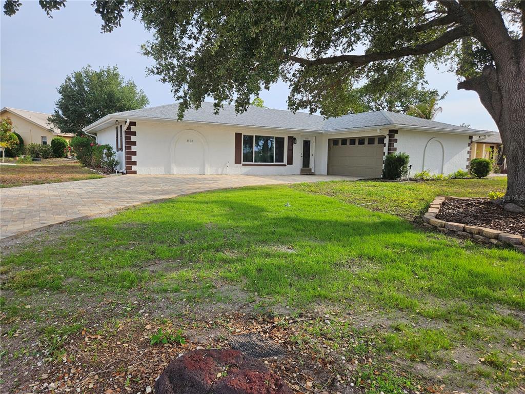 A single-story white house features a paved driveway, a two-car garage, and a large tree — 1508 FLAMINGO LANE, SUN CITY CENTER, FL