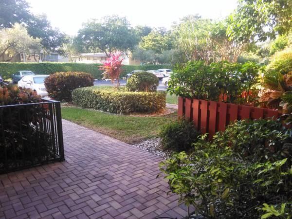 A brick patio leads to a landscaped yard with a red fence and parking area — 370 NW 76th Avenue # 104, Margate FL 33063, MARGATE, FL