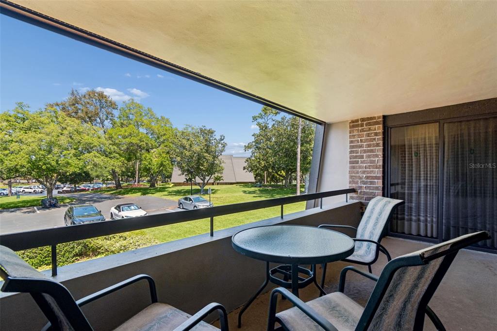 A balcony with a small table and chairs overlooks a parking lot and green space — 36750 US HIGHWAY 19 N UNIT 22208, PALM HARBOR, FL