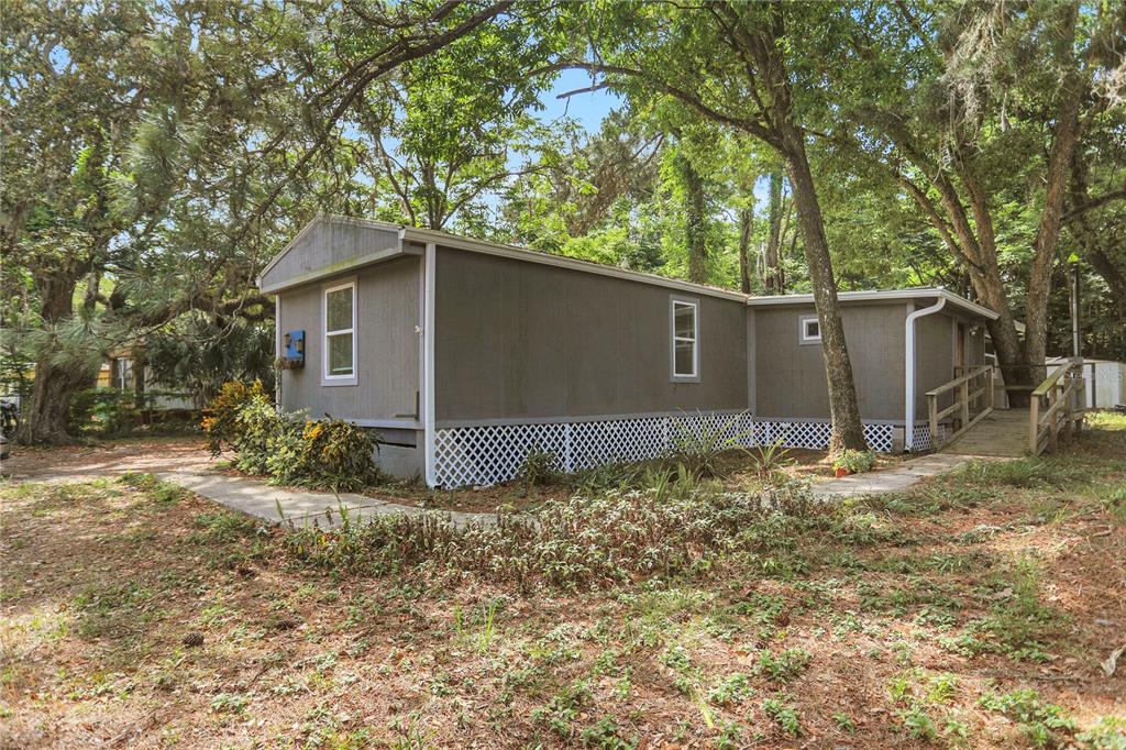 A gray mobile home with white lattice skirting sits in a wooded yard with trees — 9132 MANGO STREET, NEW PORT RICHEY, FL