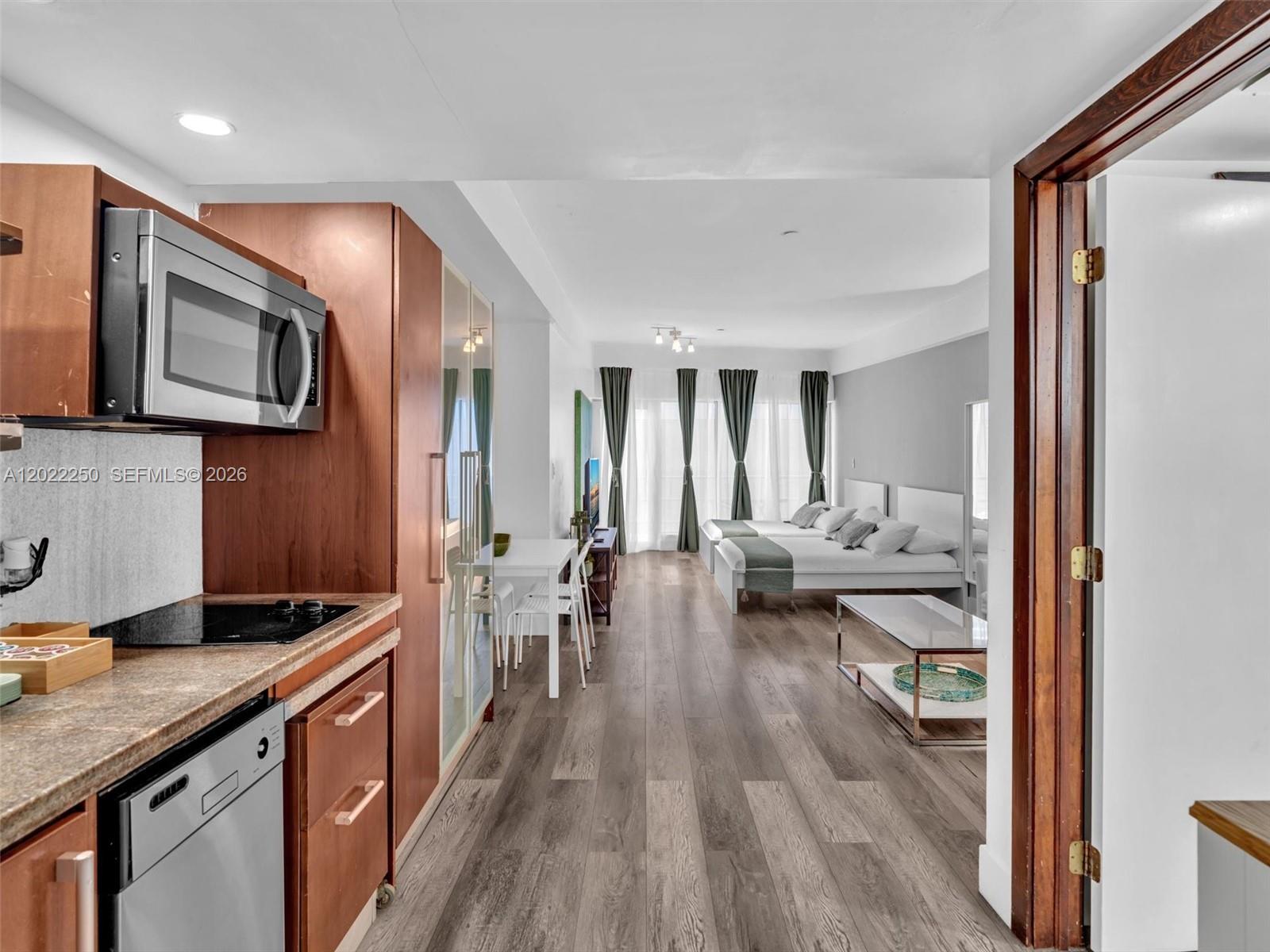 A kitchen with wood cabinets opens into a bedroom area with two beds and curtains — 1052 Ocean Dr # C203, Miami Beach FL 33139, MIAMI BEACH, FL