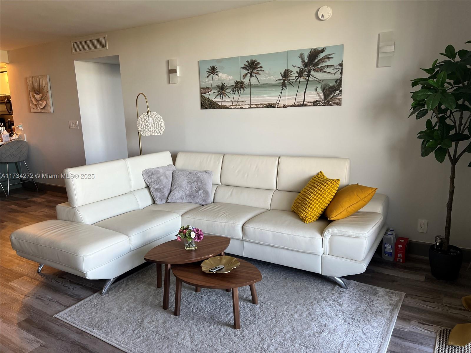 A living room features a white leather sectional sofa, nesting coffee tables, and palm tree artwork — 600 Three Islands Blvd # 504, Hallandale Beach FL 33009, HALLANDALE BEACH, FL