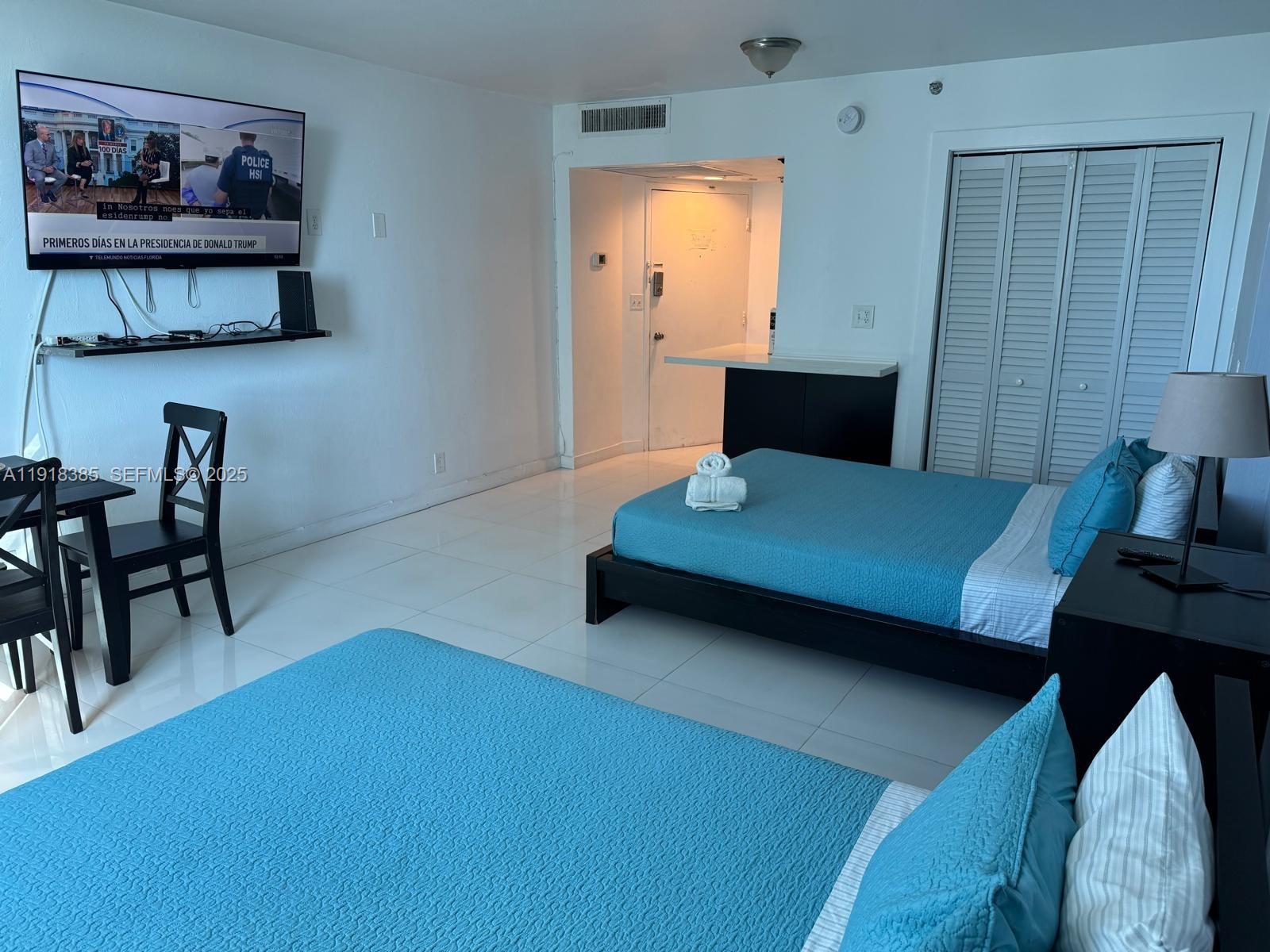 A hotel room features two beds with blue bedding, a television, and a small dining area — 5445 Collins Ave # 915, Miami Beach FL 33140, MIAMI BEACH, FL