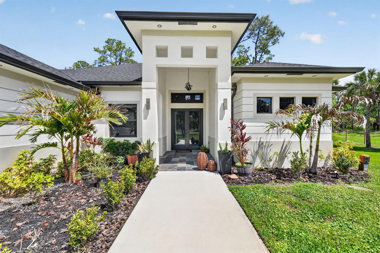 A modern house exterior features a central entryway with a concrete walkway and palm trees — 3263 SE 40th Ave Avenue SE, Naples FL 34117, NAPLES, FL