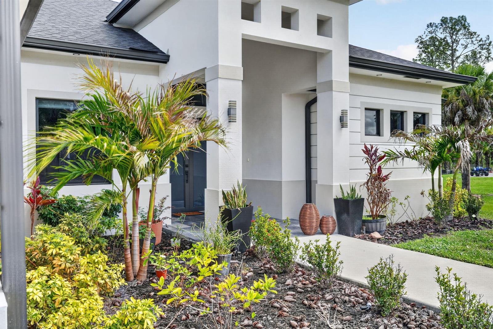 The exterior of a modern white house features a covered entryway, palm trees, and landscaping — 3263 SE 40th Ave Avenue SE, Naples FL 34117, NAPLES, FL