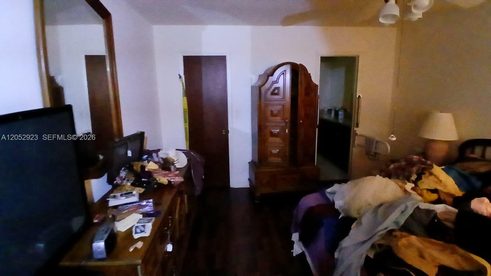 A bedroom features a wooden armoire, a dresser with a television, and a cluttered bed — CORAL SPRINGS, FL