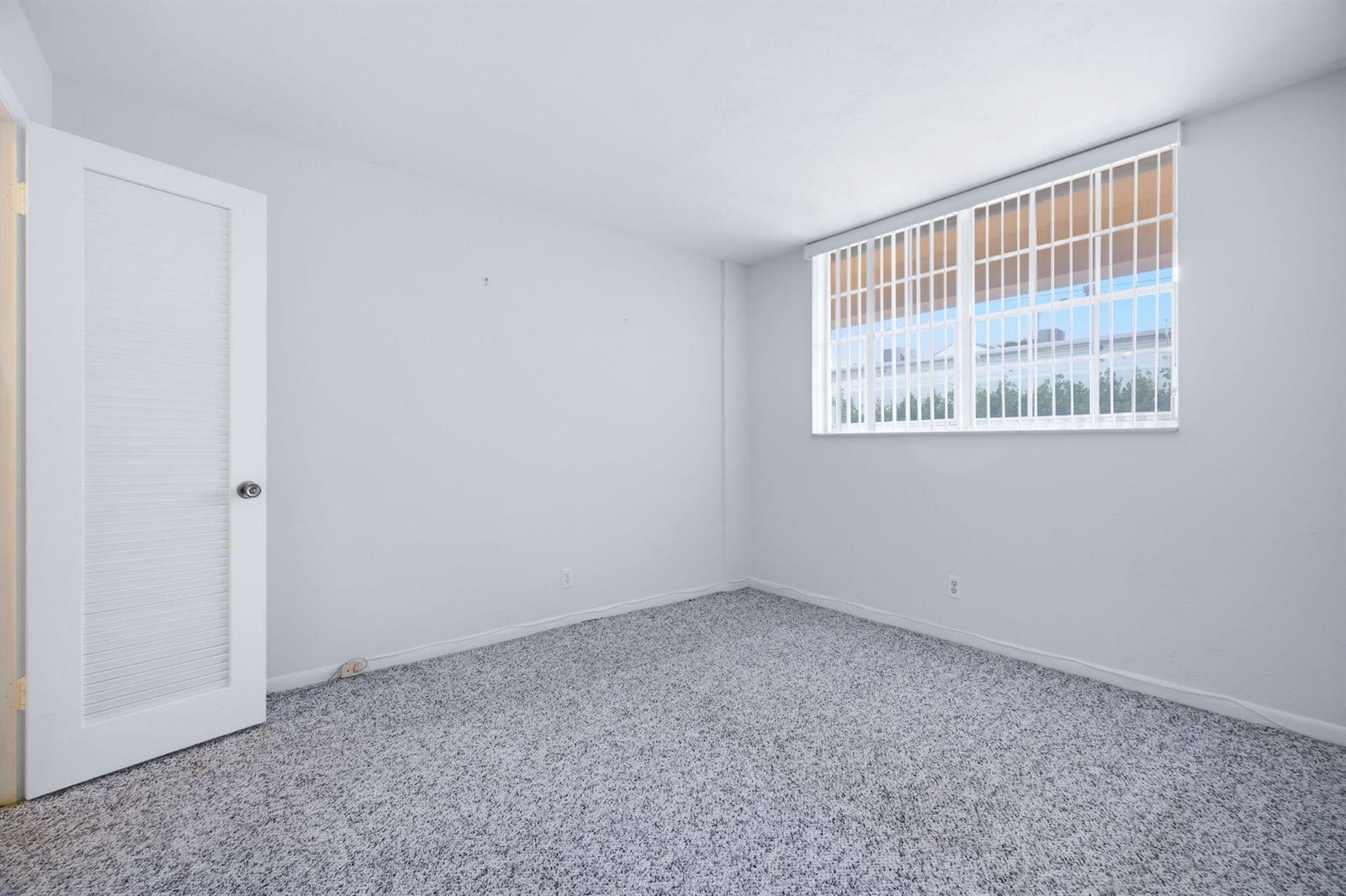 An empty bedroom features white walls, gray speckled carpeting, and a window with security bars — 4502 N Federal Highway # 125C, Lighthouse Point FL 33064, LIGHTHOUSE POINT, FL