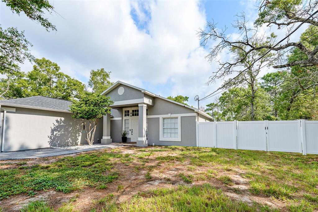 A gray single-story house features a covered entryway, a white vinyl fence, and mature trees — 11225 FLOWER AVENUE, WEEKI WACHEE, FL