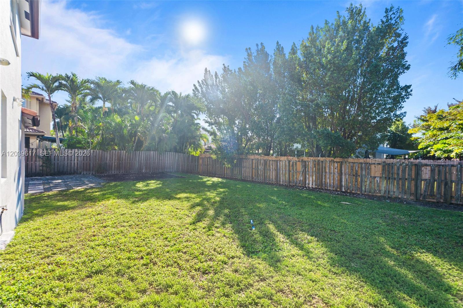 A grassy backyard features a wooden fence, mature trees, and a small paved patio area — 11558 NW 84th  St, Doral FL 33178, DORAL, FL