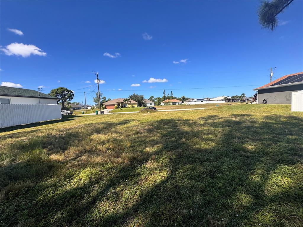 A grassy, vacant residential lot sits between two houses under a bright blue sky — 1211 NW 21ST AVENUE, CAPE CORAL, FL