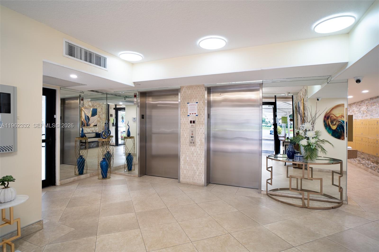 A lobby features two stainless steel elevators, tiled floors, and decorative tables with blue vases — 3510 Oaks Way # 408, Pompano Beach FL 33069, POMPANO BEACH, FL