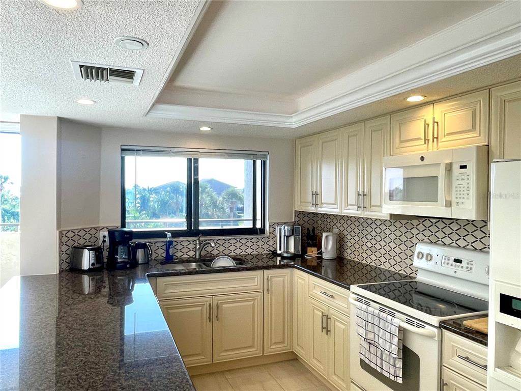 A kitchen features cream-colored cabinets, dark granite countertops, a tiled backsplash, and a window view — 1290 GULF BOULEVARD UNIT 302, CLEARWATER, FL