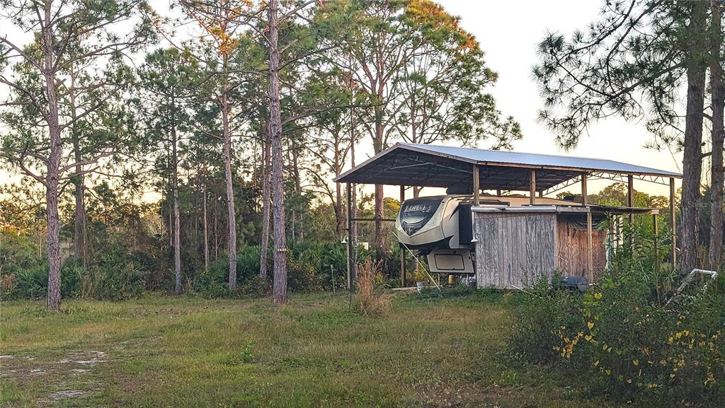 A fifth-wheel RV is parked under a metal carport structure in a wooded, grassy lot — 12885 86TH STREET, FELLSMERE, FL