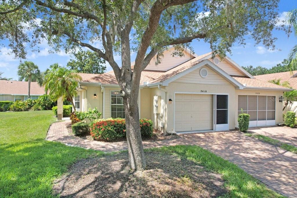 A single-story house with a beige exterior, a brick driveway, and a large oak tree — 2418 NEW HAVEN CIRCLE UNIT 83, SUN CITY CENTER, FL