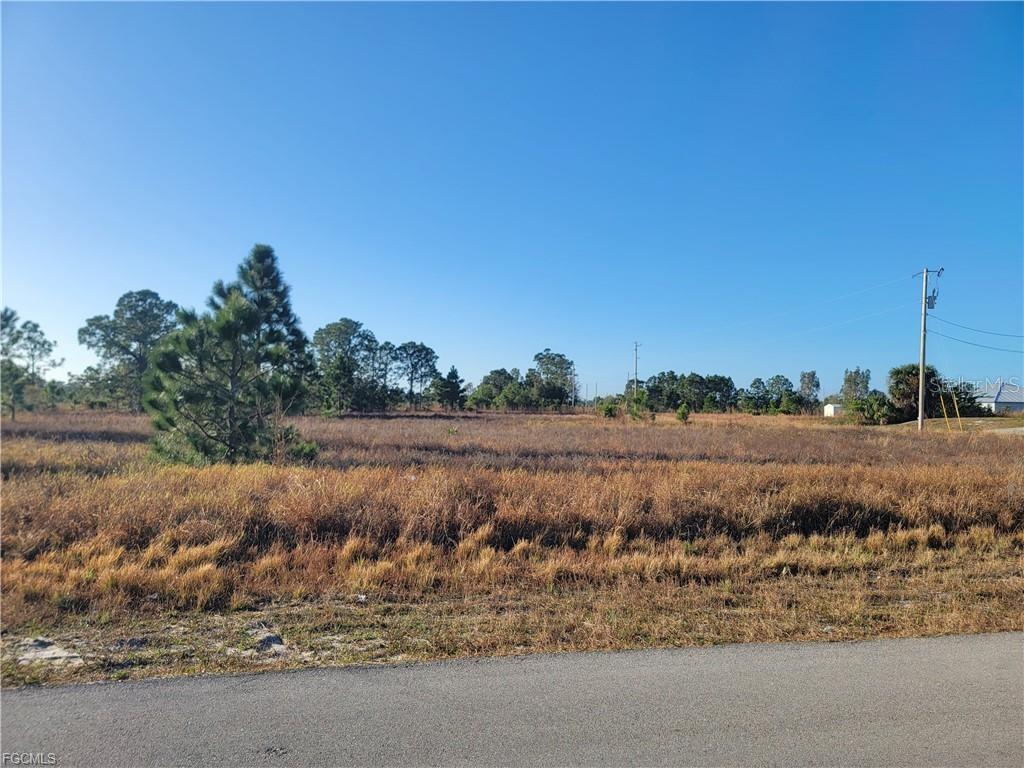A flat, grassy vacant lot with scattered trees under a clear blue sky — 707 FRONTENAC AVENUE S, LEHIGH ACRES, FL