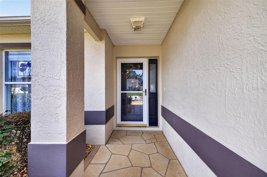 A covered entryway features a stone-tiled floor, a storm door, and beige stucco walls — 1019 EAGLES LANDING, LEESBURG, FL