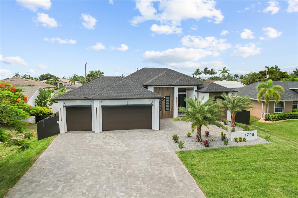 A modern single-story house with a paver driveway, three-car garage, and tropical landscaping — 1709 SW 43RD LANE, CAPE CORAL, FL