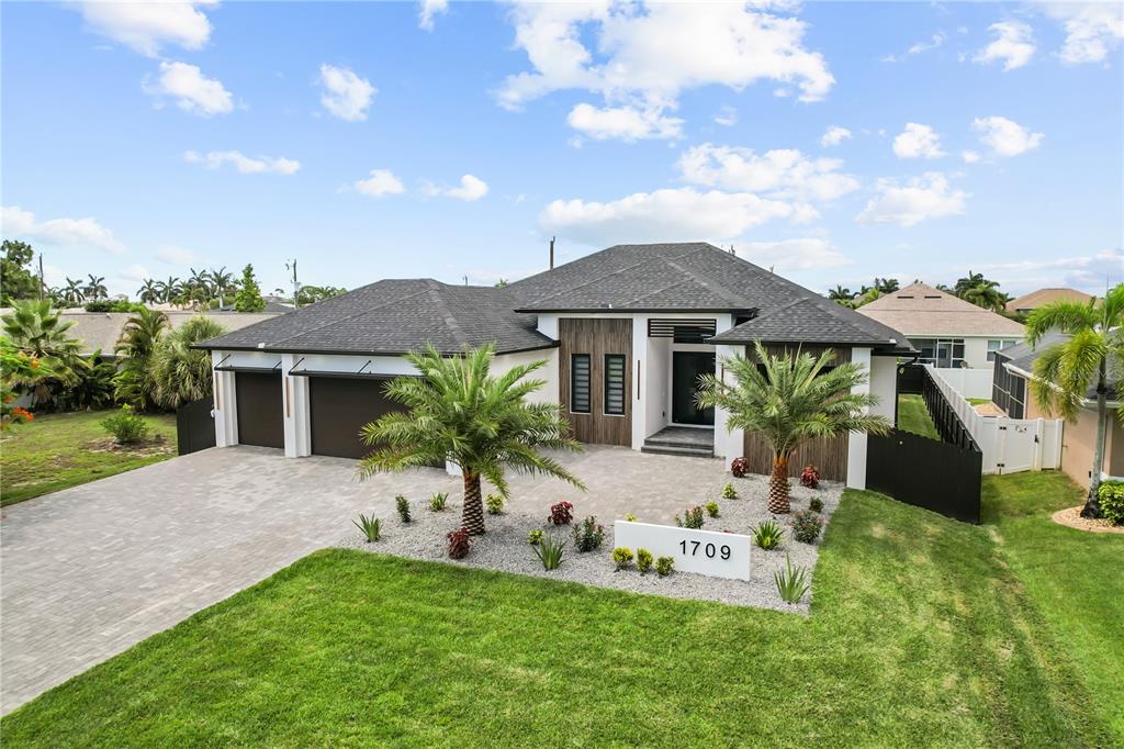 A modern single-story house with a gray shingled roof, dark garage doors, and palm trees — 1709 SW 43RD LANE, CAPE CORAL, FL