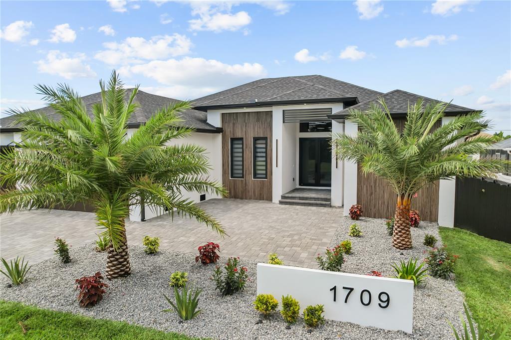 A modern single-story house features a paved driveway, two palm trees, and wood-paneled accents — 1709 SW 43RD LANE, CAPE CORAL, FL