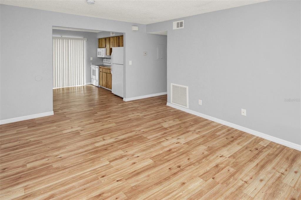An empty living room with light wood-look flooring leads into a kitchen with white appliances — 7450 35TH STREET N UNIT 1604, PINELLAS PARK, FL