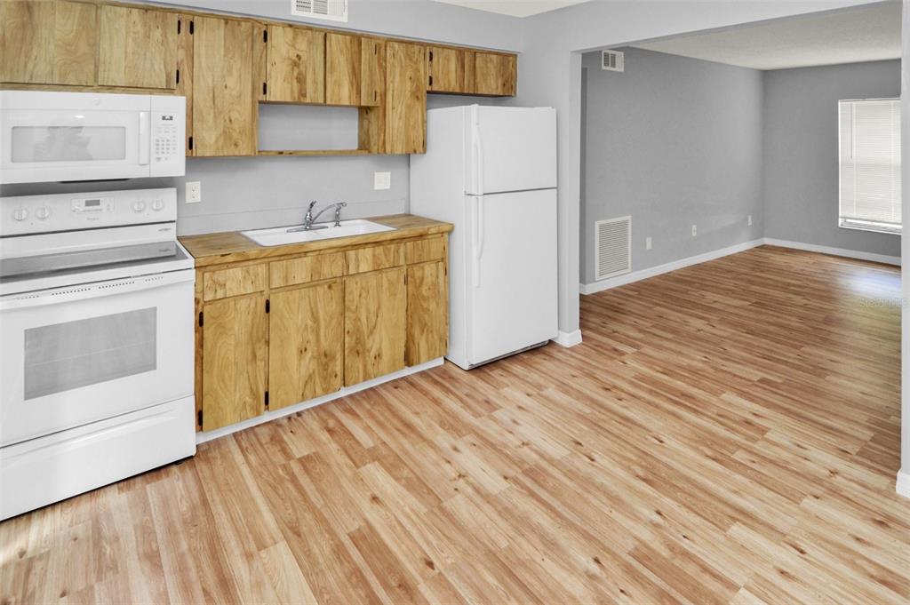 A kitchen with wood cabinets, white appliances, and light wood-look flooring opens to a living area — 7450 35TH STREET N UNIT 1604, PINELLAS PARK, FL