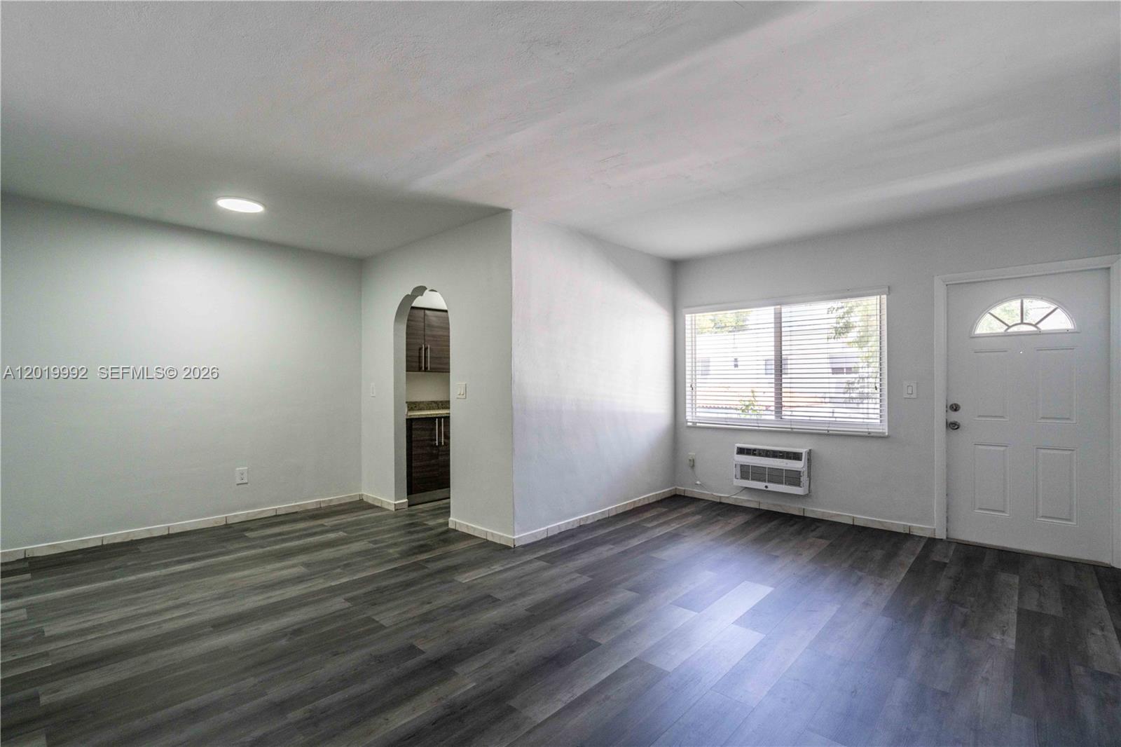 An empty living room features gray wood-look flooring, a window with blinds, and an arched doorway — 7911 Abbott Ave # 9, Miami Beach FL 33141, MIAMI BEACH, FL