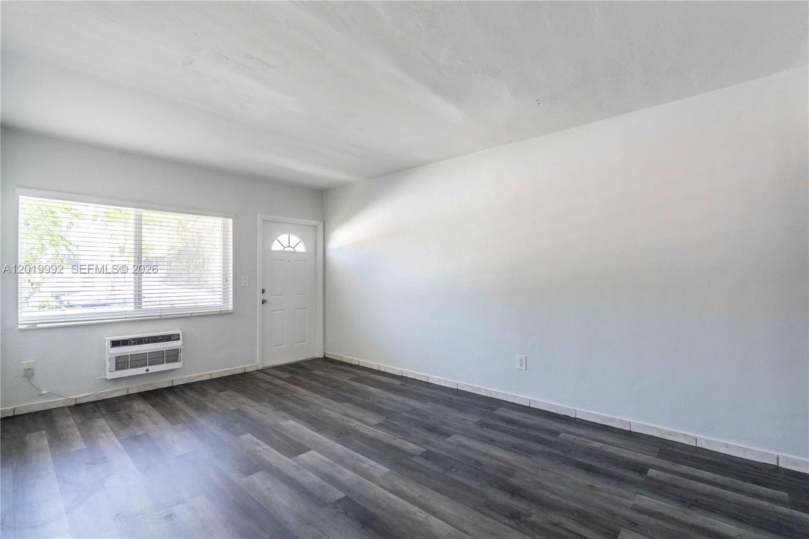An empty room features light gray walls, dark wood-look flooring, a window, and a door — 7911 Abbott Ave # 9, Miami Beach FL 33141, MIAMI BEACH, FL