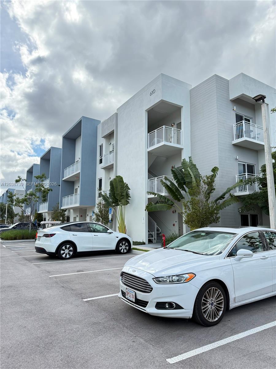 A white multi-story apartment building with balconies is shown behind two parked cars — 610 NE 2nd St # 301, Dania Beach FL 33004, DANIA BEACH, FL