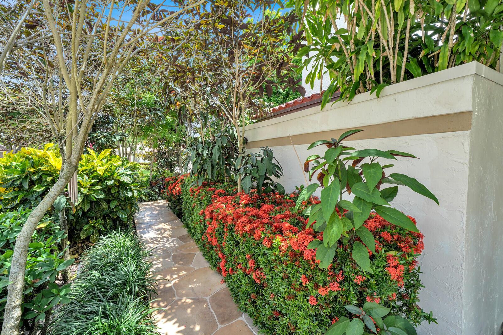 A stone pathway leads past a vibrant red ixora hedge and lush landscaping beside a wall — 21381 Sonesta Way, Boca Raton FL 33433, BOCA RATON, FL