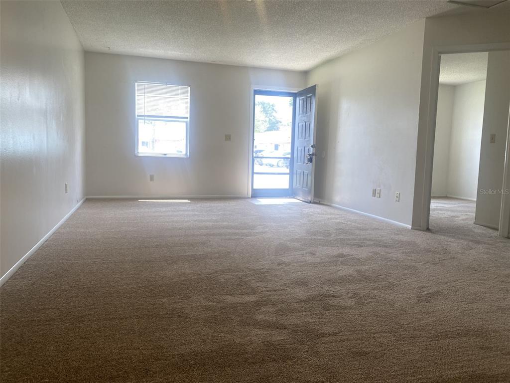 An empty living room features beige carpeting, white walls, a window, and an open front door — 47 W LAS PALMAS WAY, KISSIMMEE, FL