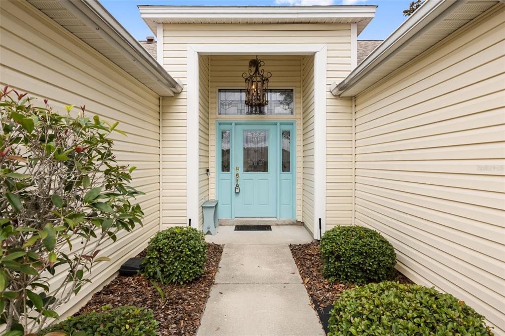 A light blue front door with glass panels sits under a covered entryway with siding — 1312 BAEZ WAY, THE VILLAGES, FL